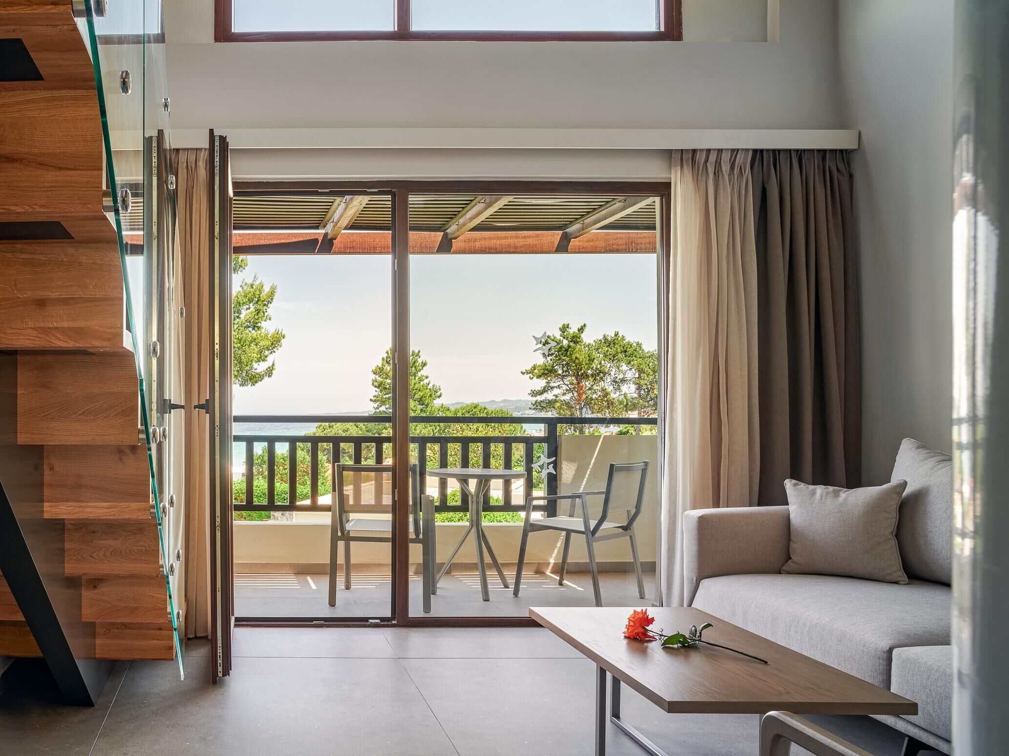 A modern living room with a sofa, a coffee table, and a balcony with a view of greenery and a body of water.