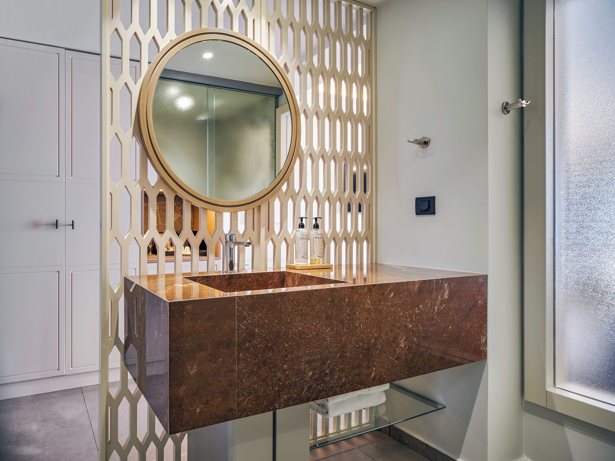 Un baño con lavamanos de mármol, un espejo redondo y un separador decorativo.