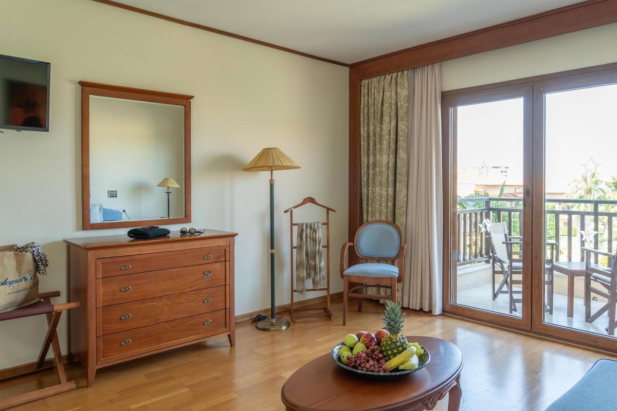 Une chambre d’hôtel avec une commode en bois, un miroir, une lampe, une chaise, une table avec des fruits et un balcon aménagé avec du mobilier d’extérieur.
