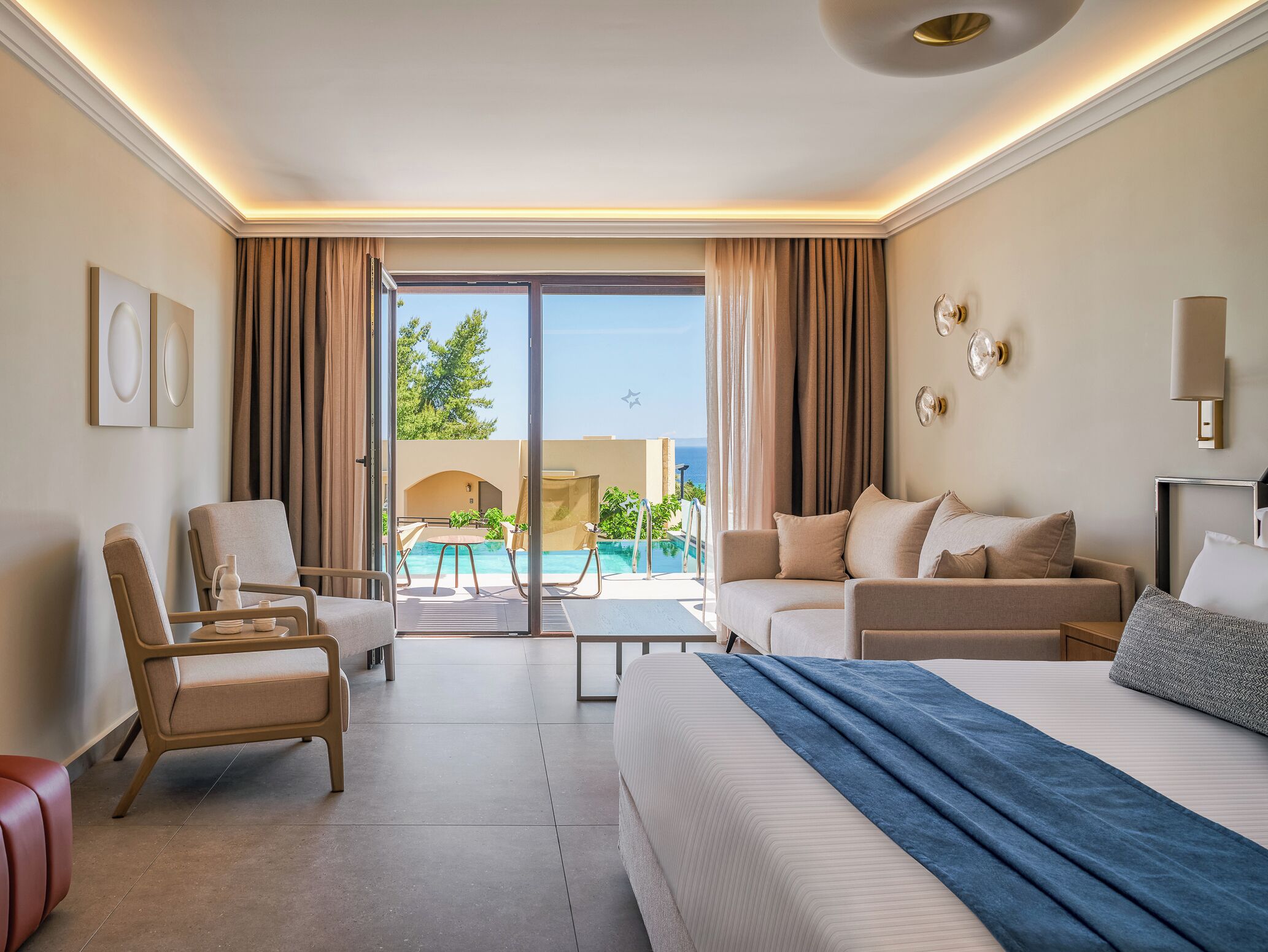 A hotel room with a bed, two armchairs, a sofa, a small table, and a view of a pool and ocean.