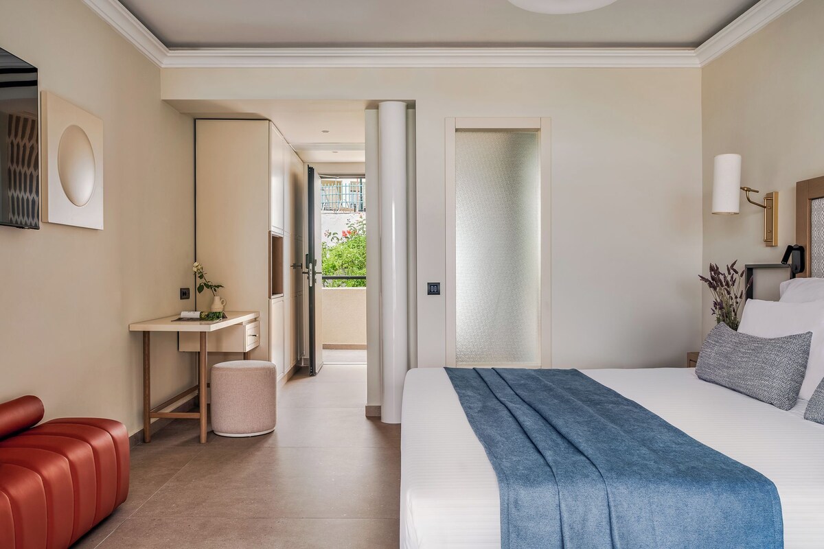 A hotel room with a bed, a red leather sofa, a small table with a plant, and a view of the outside balcony.