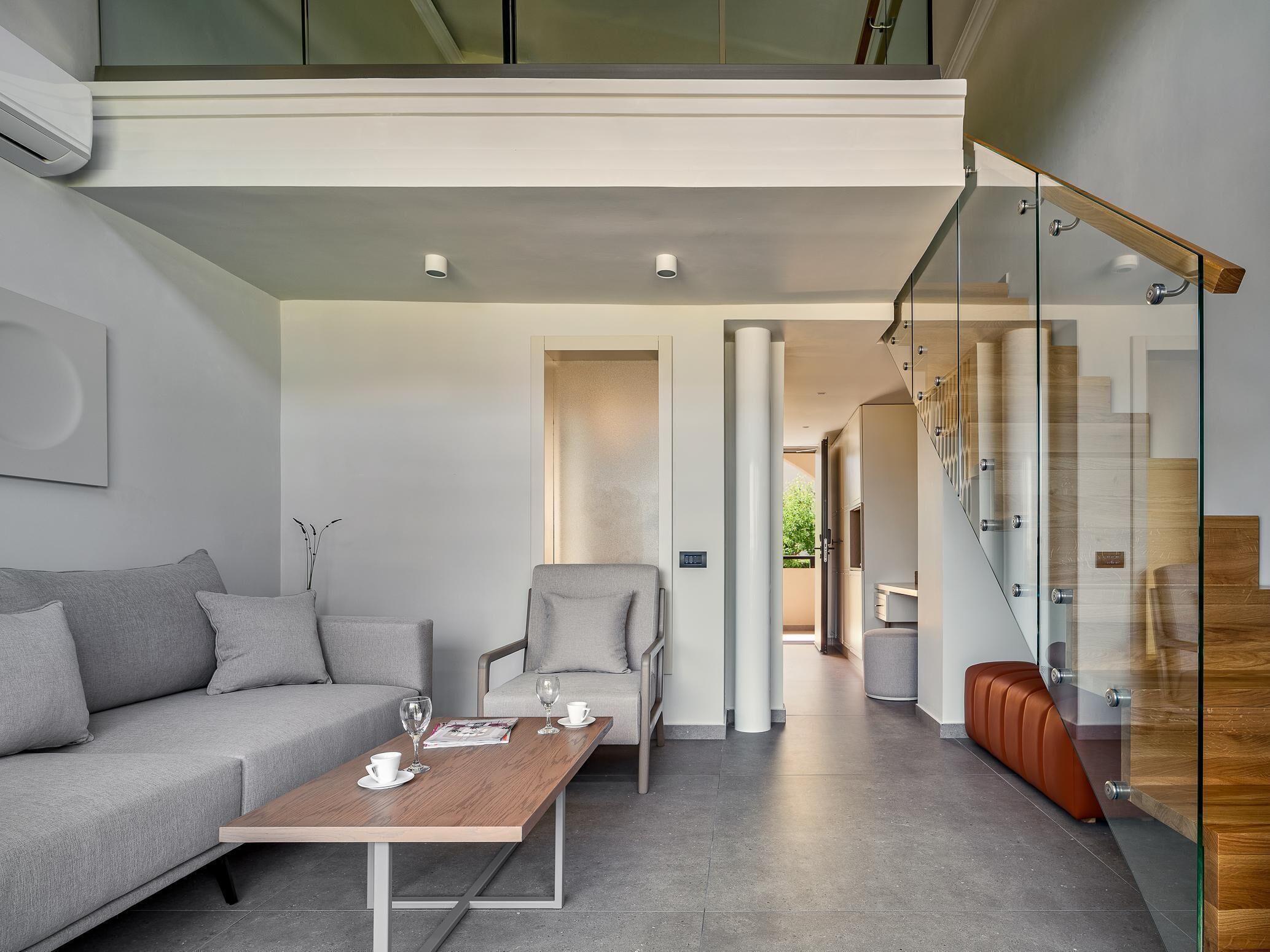 A modern living room with a sofa, armchair, and coffee table. A staircase with glass railings leads to an upper level.