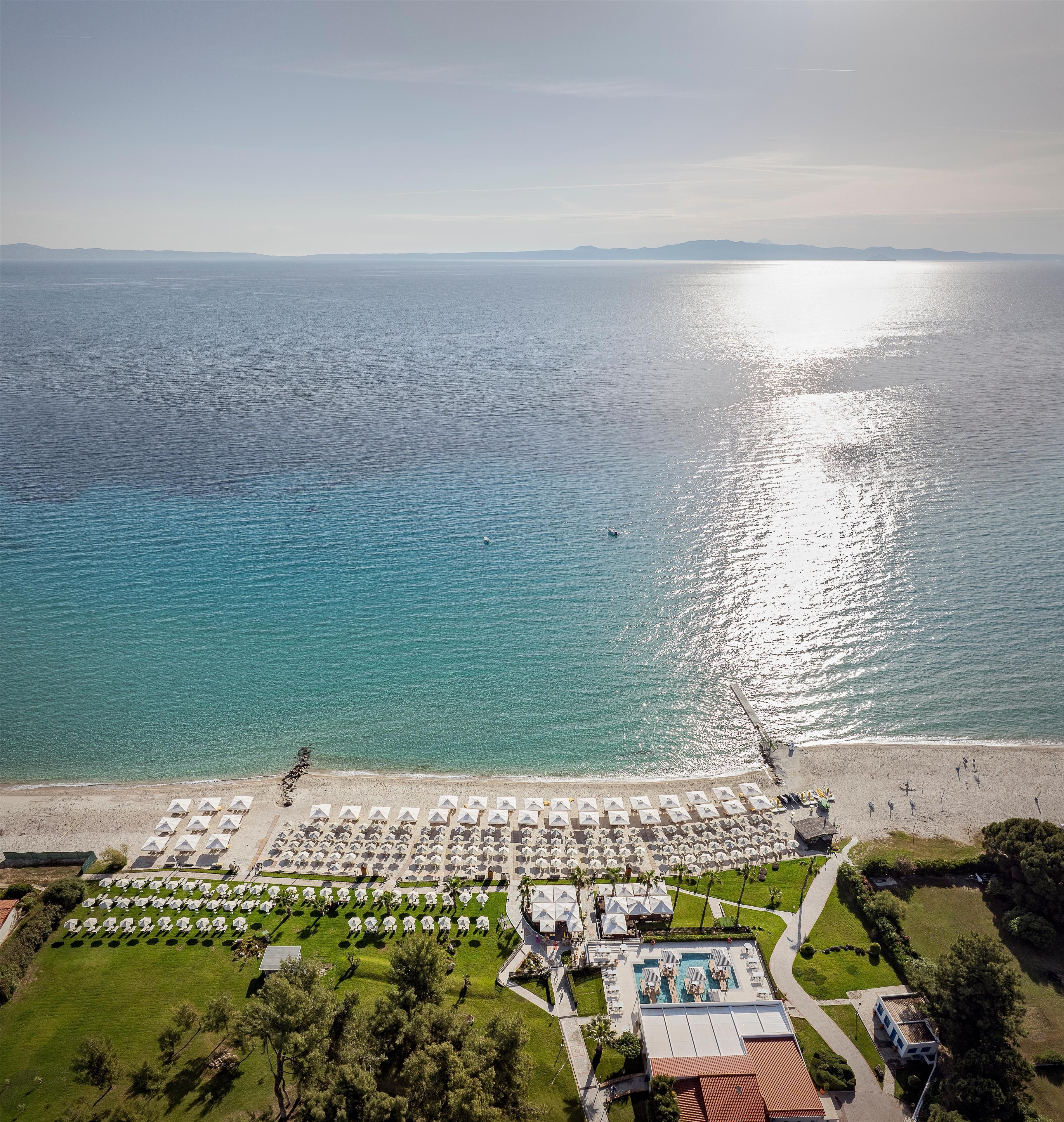 An aerial view of a beach resort with rows of beach umbrellas, a swimming pool, and a clear view of the sea.