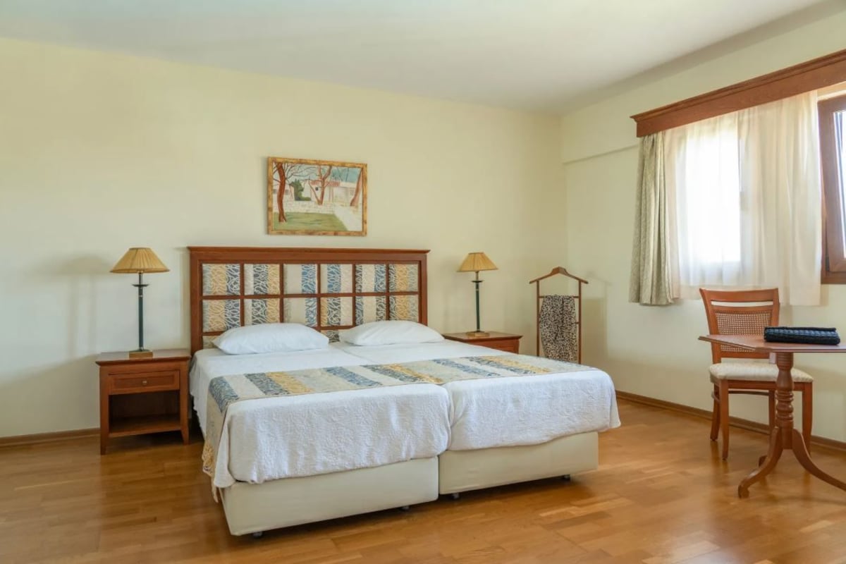 A double bed with a patterned headboard, two bedside tables with lamps, a chair, and a window with curtains.