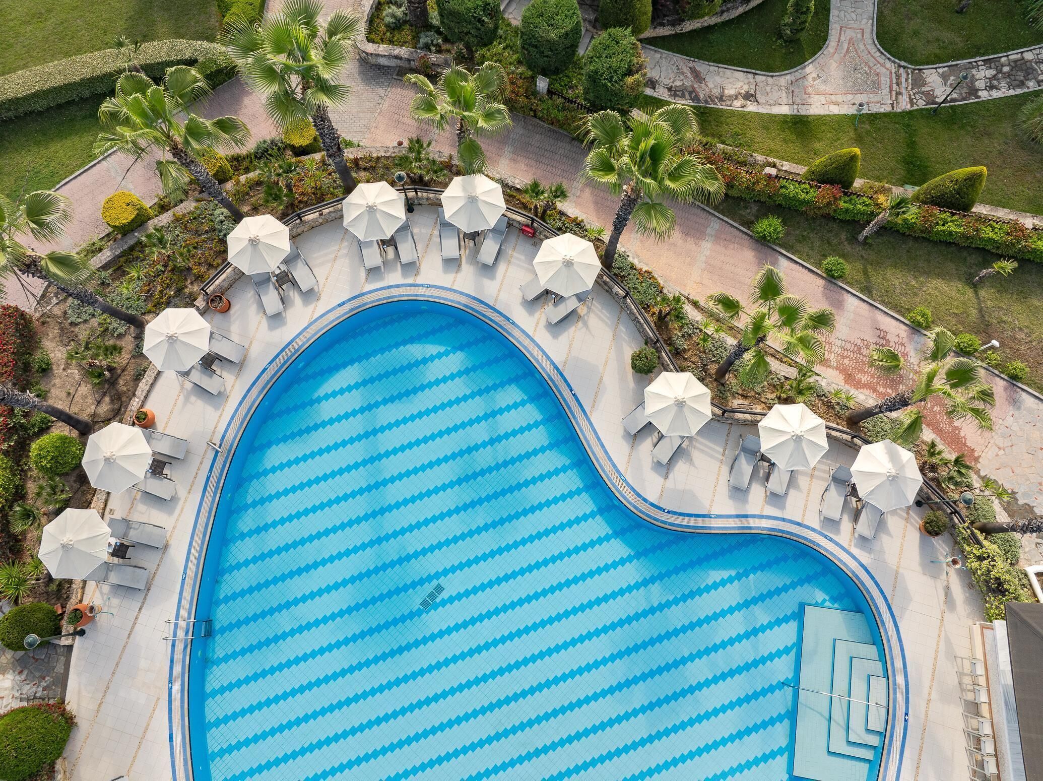 An aerial view of a swimming pool with a zigzag pattern, surrounded by white umbrellas and lounge chairs, with palm trees and landscaped gardens.