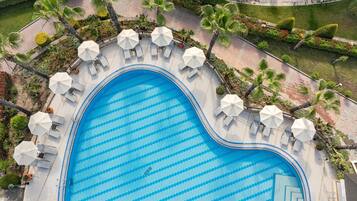 An aerial view of a swimming pool with a zigzag pattern, surrounded by white umbrellas and lounge chairs, with palm trees and landscaped gardens.