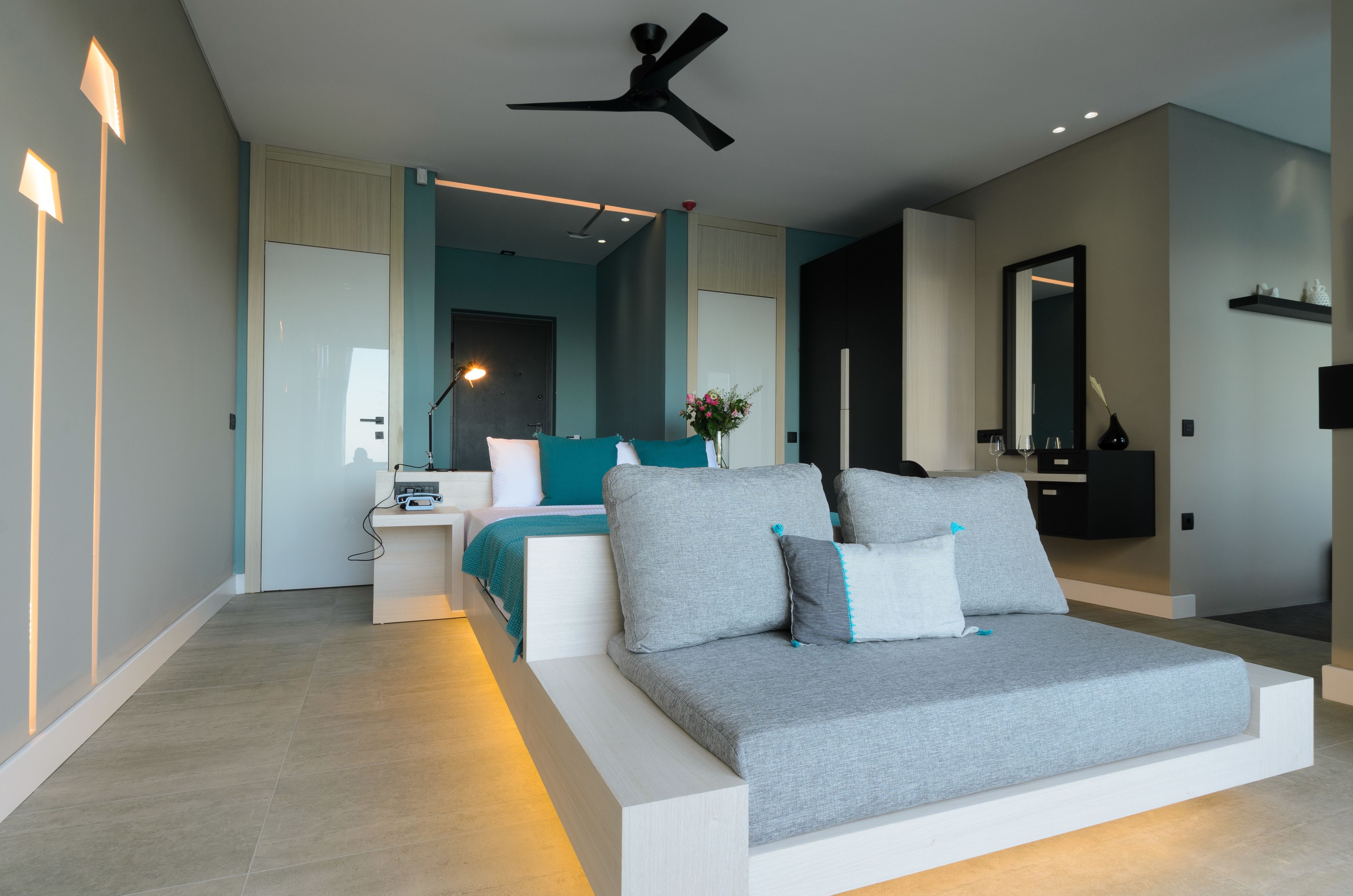 Une chambre moderne avec un canapé gris, un lit avec une literie de couleur turquoise, une table de chevet avec une lampe et un ventilateur de plafond.