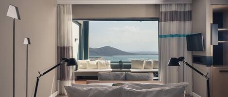 A modern hotel room with a bed, two bedside lamps, a TV, and a view of the sea and mountains.