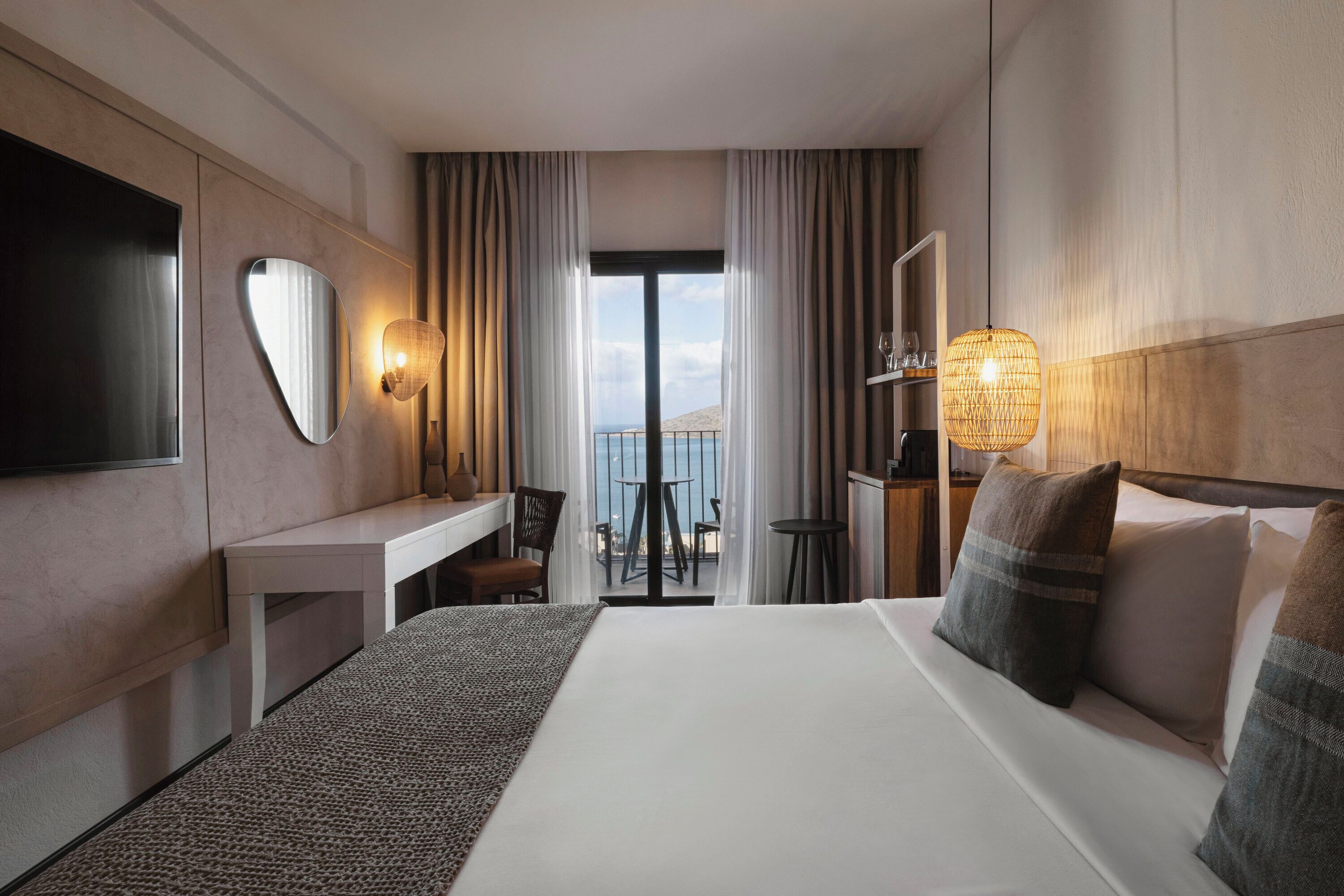 A hotel room with a large bed, a desk, a TV, and a balcony with a view of the sea.
