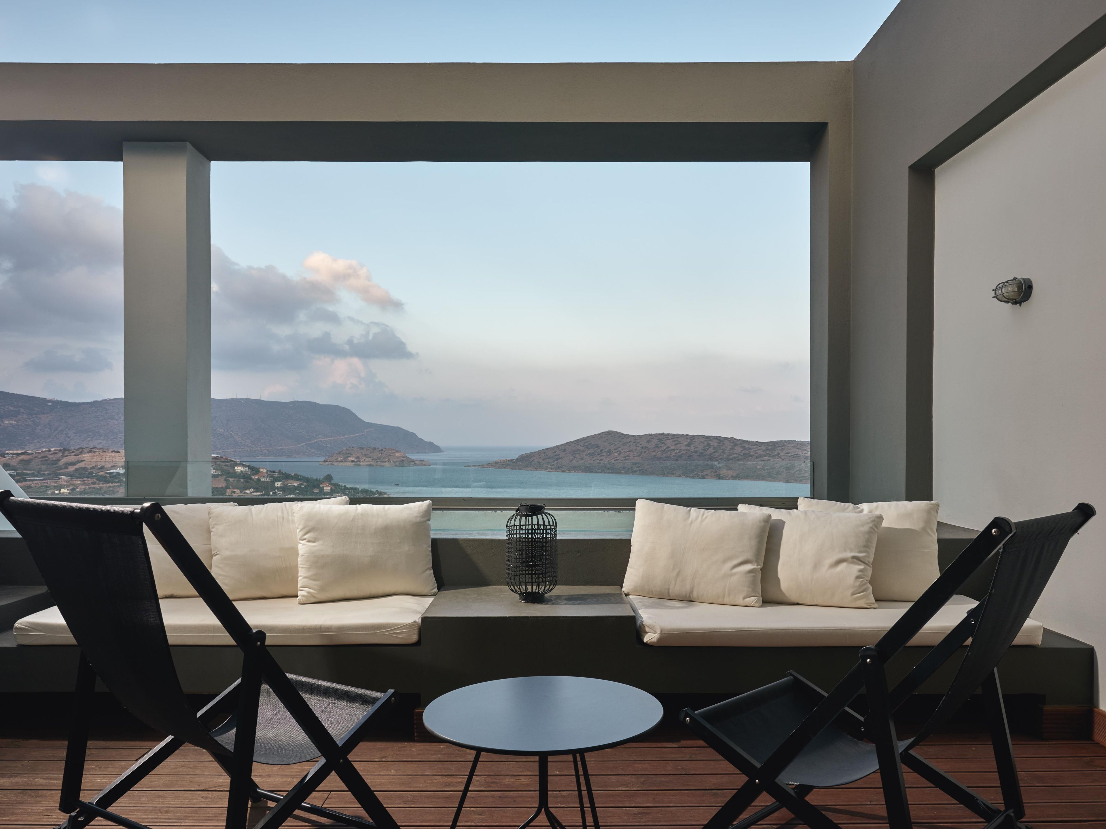 A balcony with a sofa, chairs, and a small table, overlooking a scenic view of hills and a body of water.