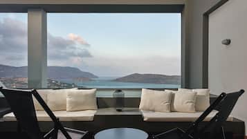 A balcony with a sofa, chairs, and a small table, overlooking a scenic view of hills and a body of water.