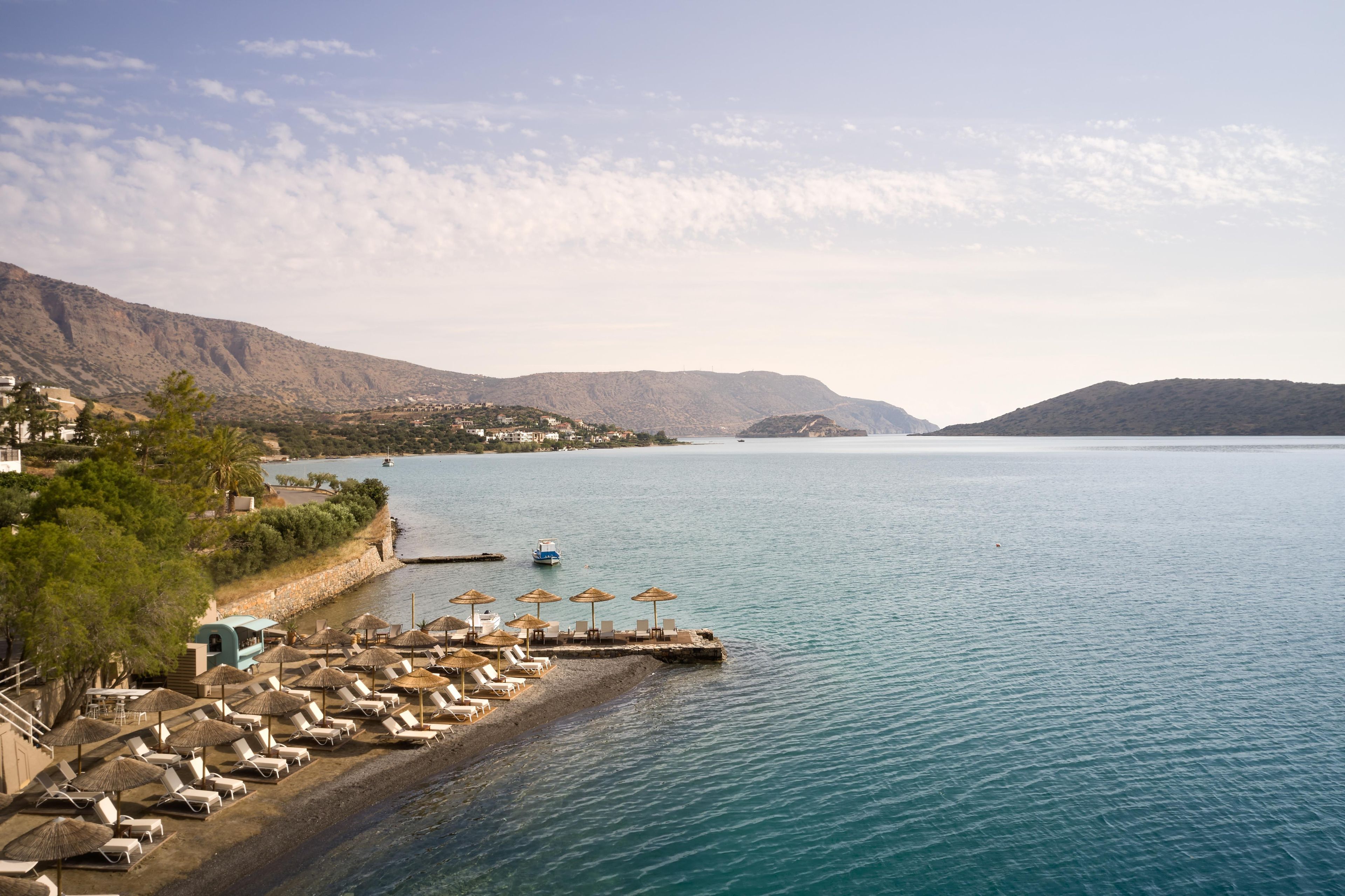A coastal resort with a beach, lounge chairs, umbrellas, and a view of the sea and mountains.