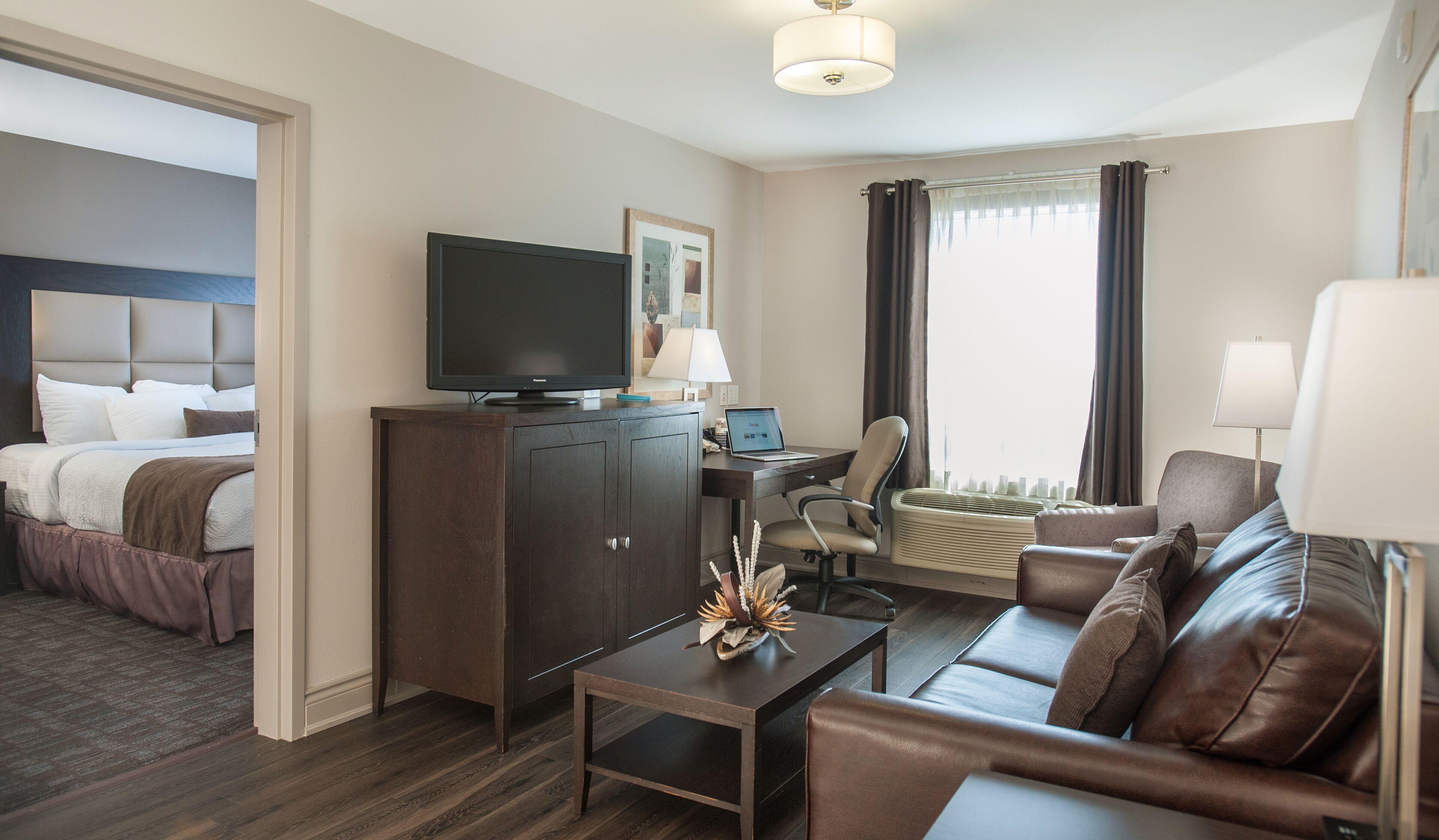 A hotel room with a bed, a desk with a laptop, a television, a sofa, and a window with curtains.