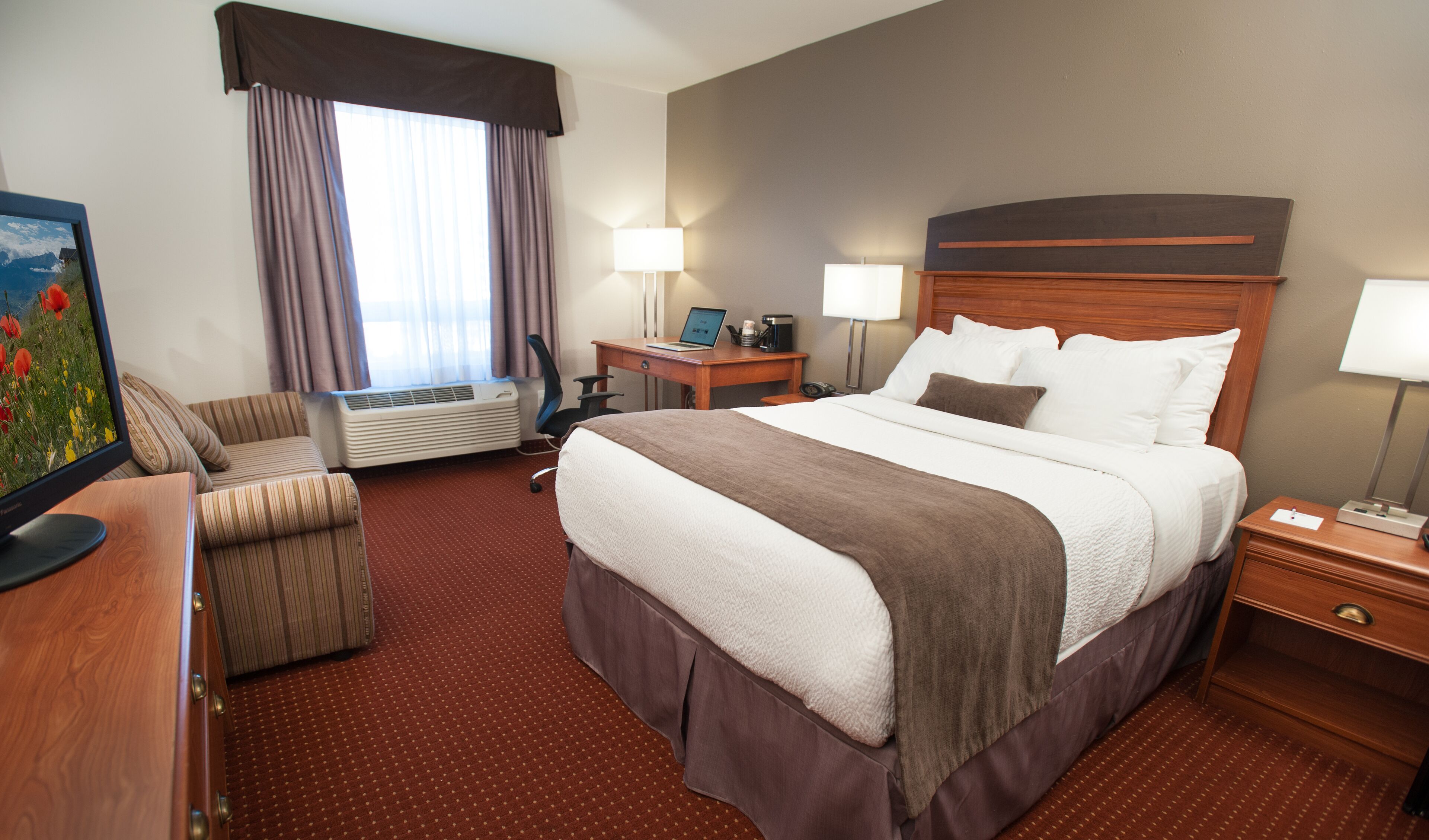 A hotel room with a large bed, a desk, a chair, a desk lamp, a bedside table, and a wall-mounted TV.