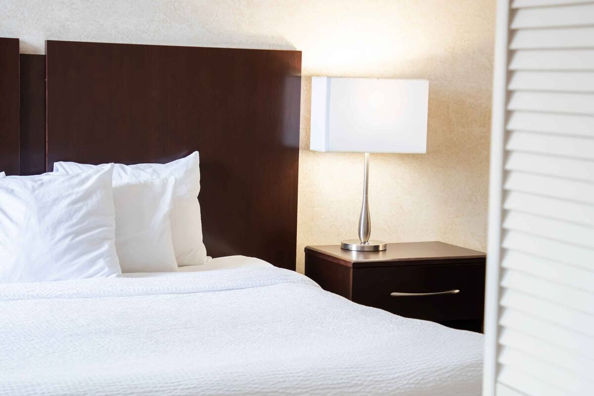 Una cama bien hecha con sábanas y almohadas blancas, una mesita de noche con lámpara y un cabecero de madera.