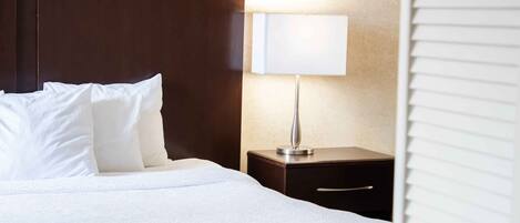 A neatly made bed with white linens and pillows, a bedside table with a lamp, and a wooden headboard.