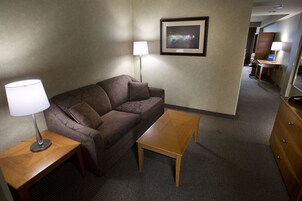 A hotel room with a brown sofa, a wooden coffee table, a lamp, a framed picture on the wall, and a desk with a chair.