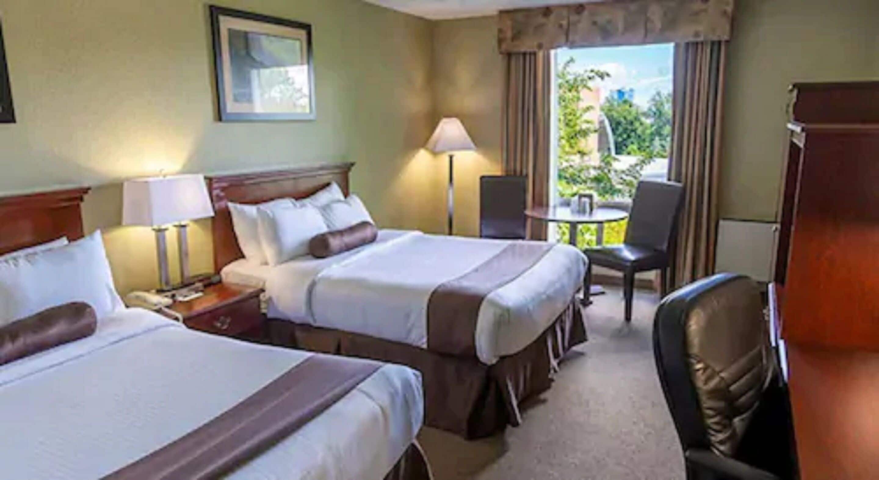 A hotel room with two beds, a desk, a chair, and a balcony with a view of greenery.