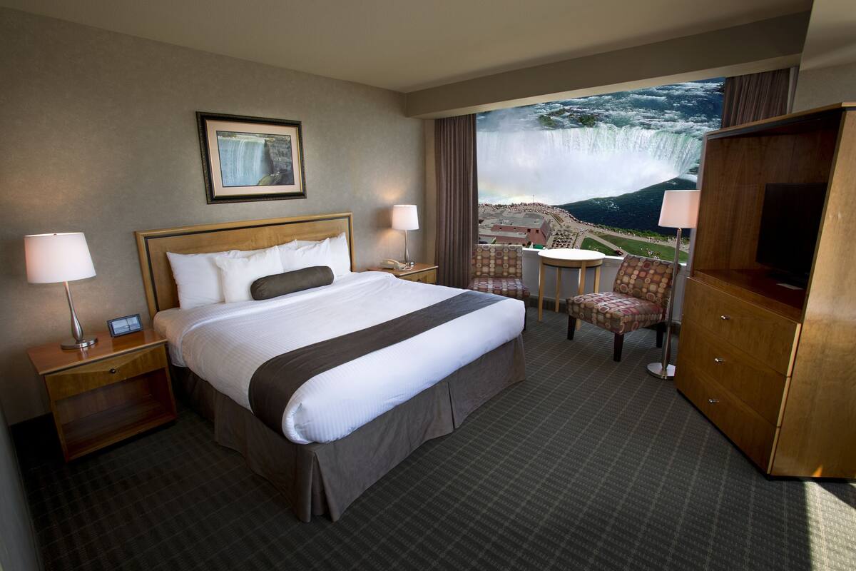 Habitación de hotel con una cama grande, televisor, escritorio y una silla. Hay un cuadro en la pared y vistas a una cascada por la ventana.