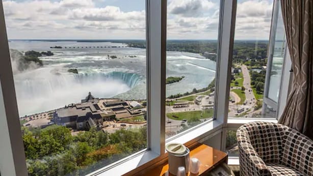 A room with a large window overlooking a waterfall and a distant cityscape.