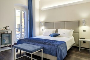 Quarto de hotel com cama, mesa de cabeceira, banquinho e um pequeno frigorífico.
