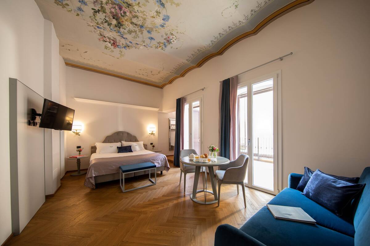 A hotel room with a bed, a desk with a chair, a blue sofa, and a TV on the wall.