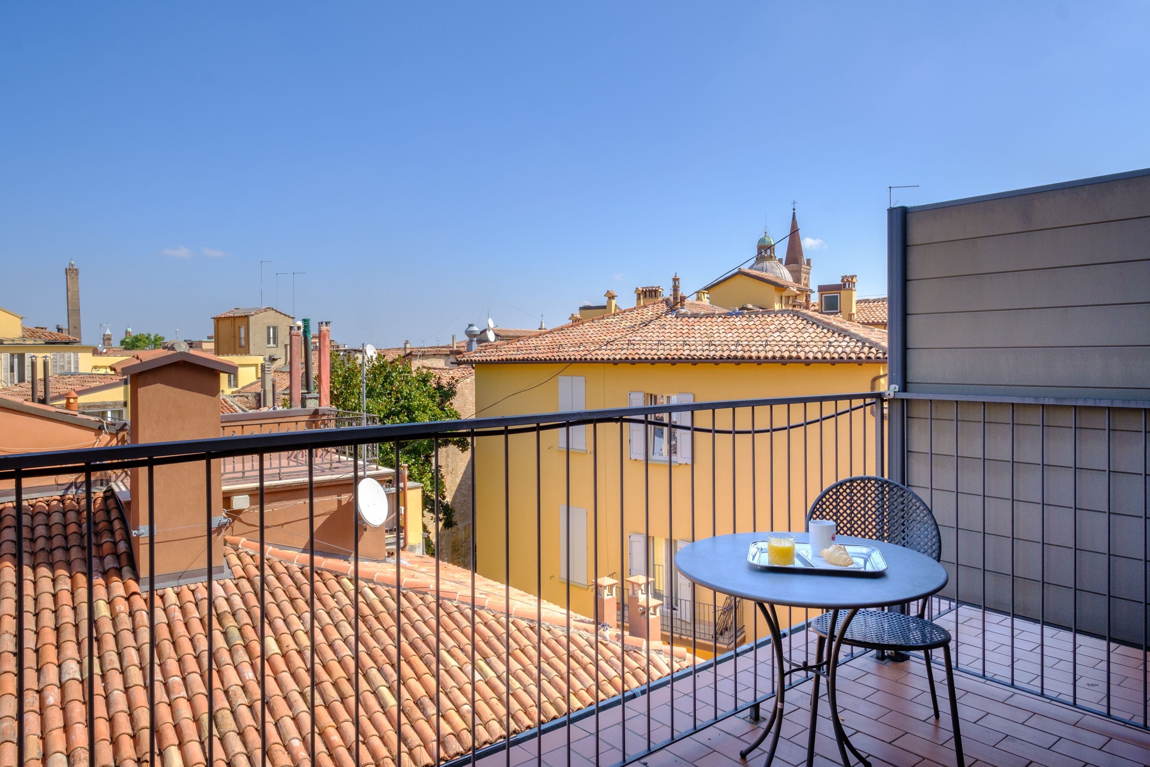 Un balcón con mesa y sillas, con vistas a un paisaje urbano de tejados de teja y una aguja de iglesia.