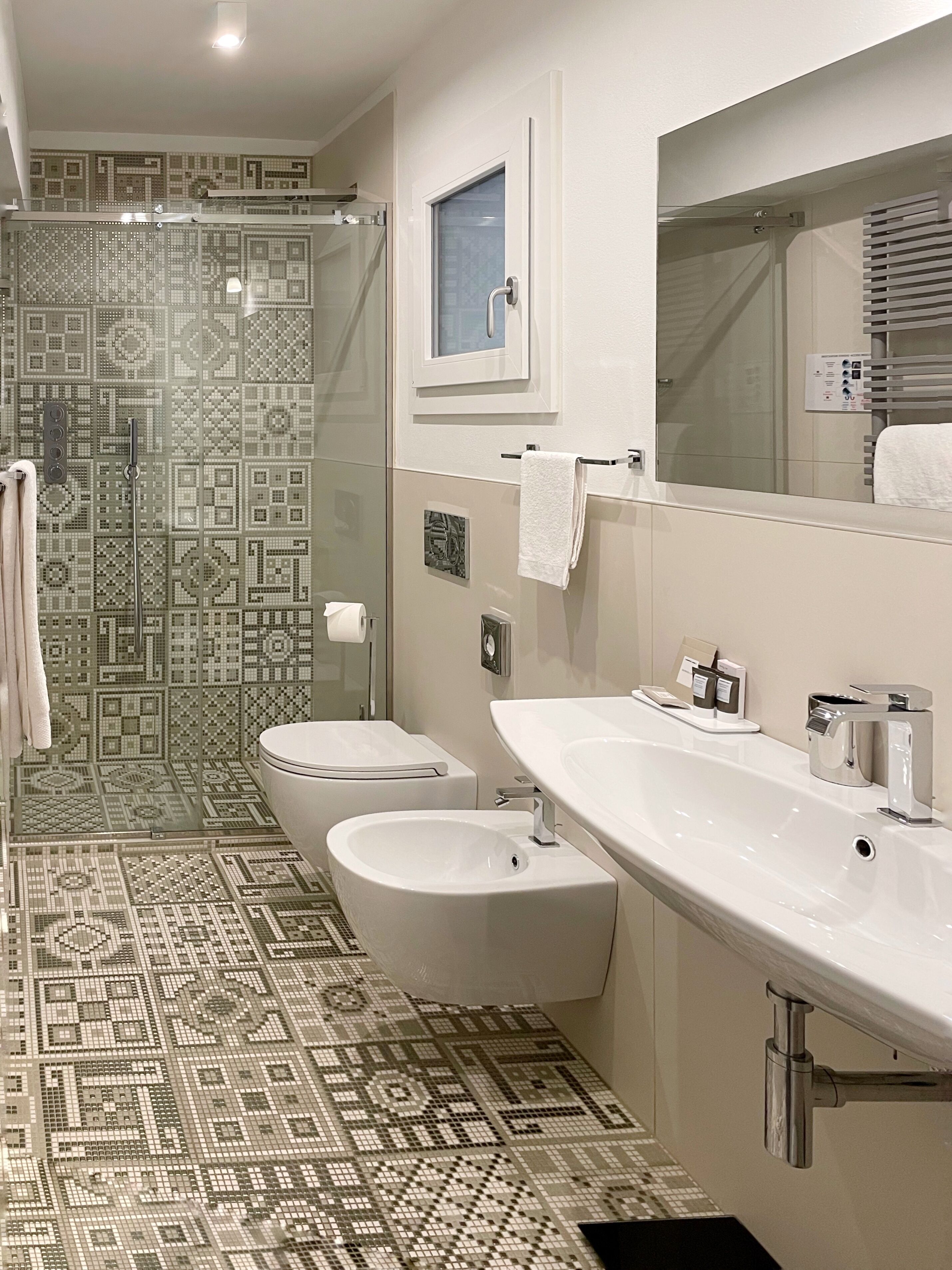 A modern bathroom with a patterned floor, two sinks, a glass shower enclosure, and a large mirror.