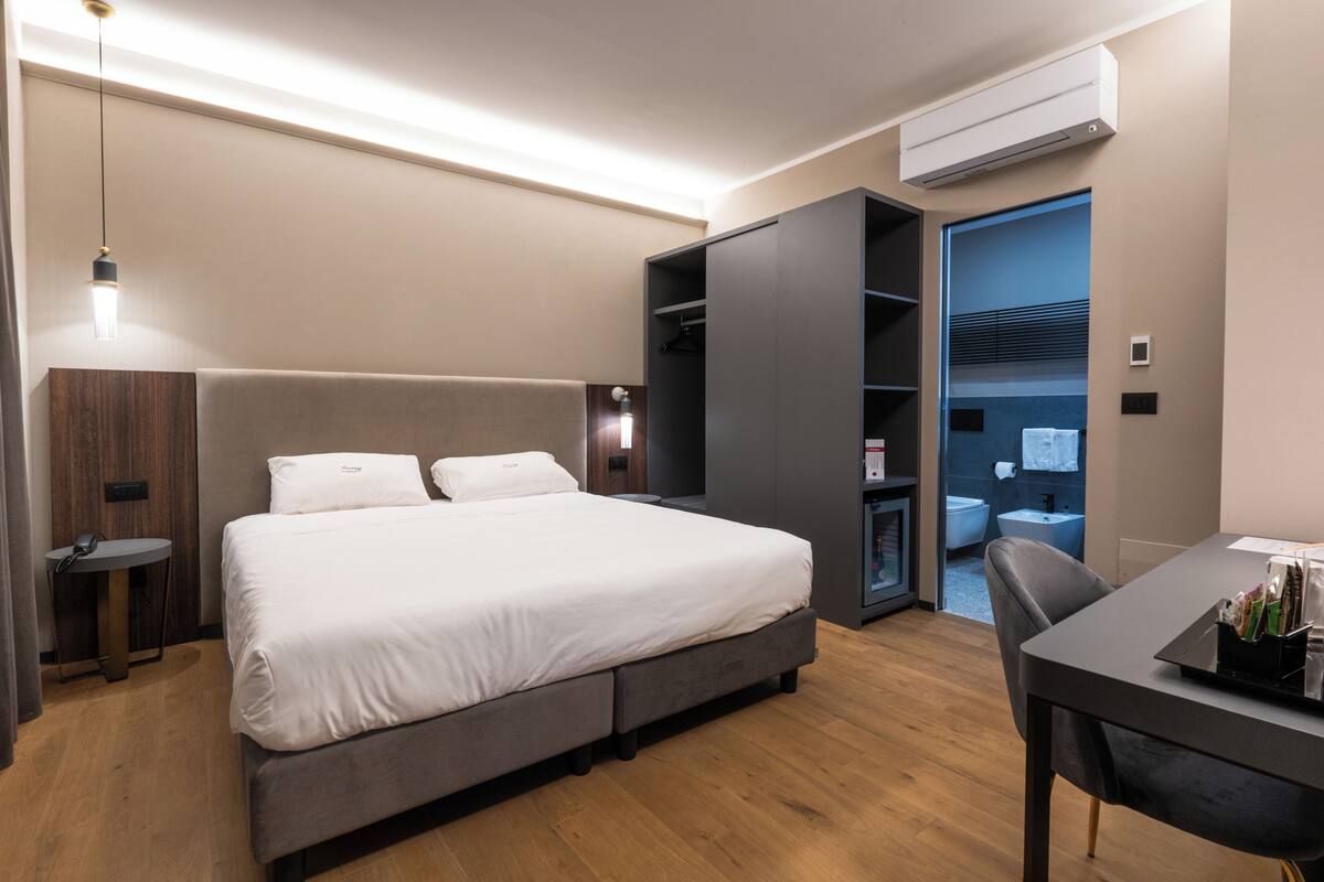 A modern hotel room with a large bed, a desk, and a wardrobe.