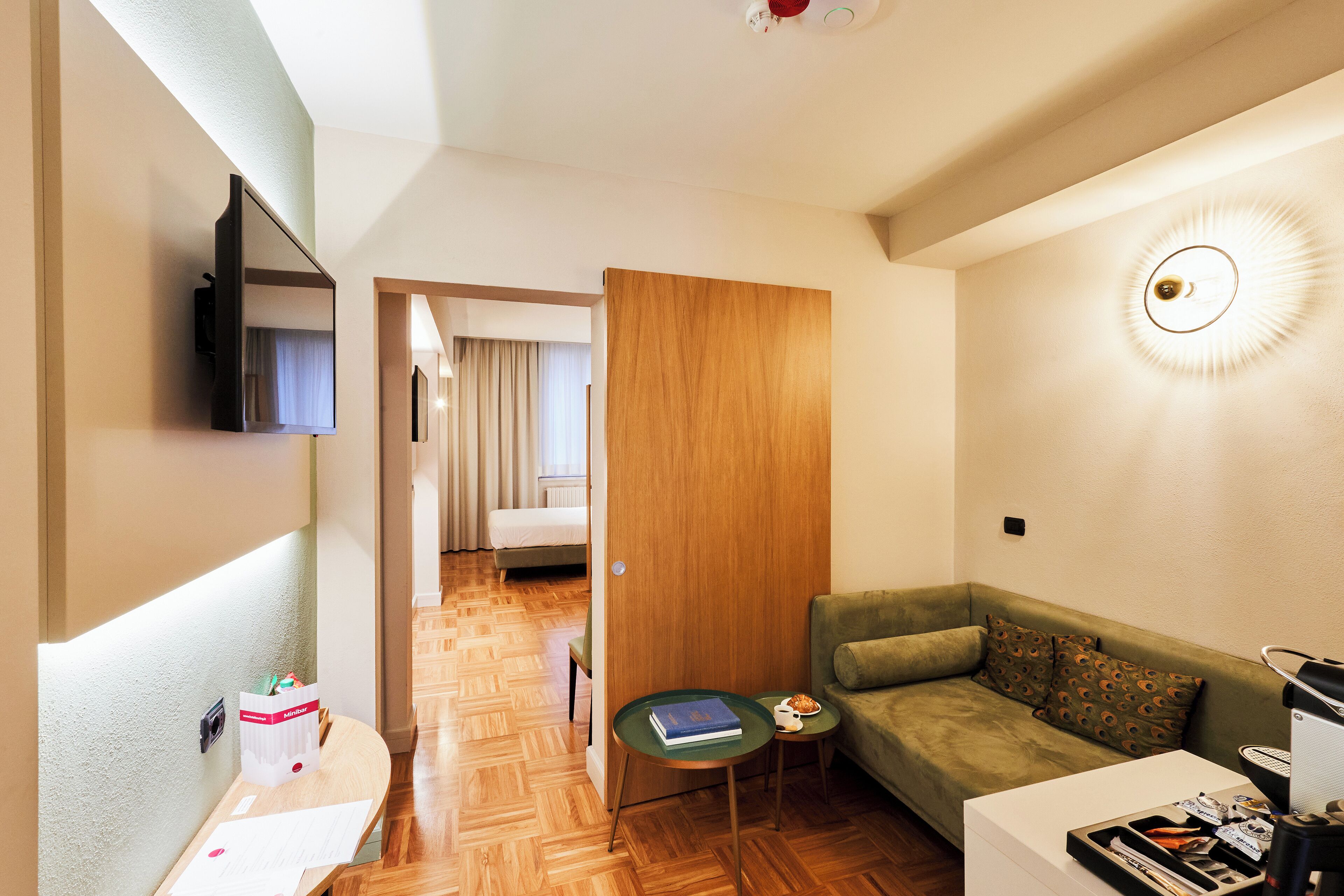 Una camera d'albergo moderna con pavimento in legno, un divano verde, un piccolo tavolino, una TV a schermo piatto montata a parete e una porta che dà su un'altra stanza.