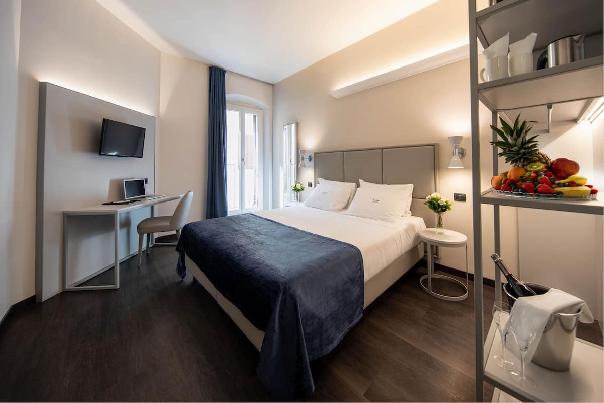 A modern hotel room with a bed, a desk with a laptop, a TV, and a small table with fruit and a bottle.