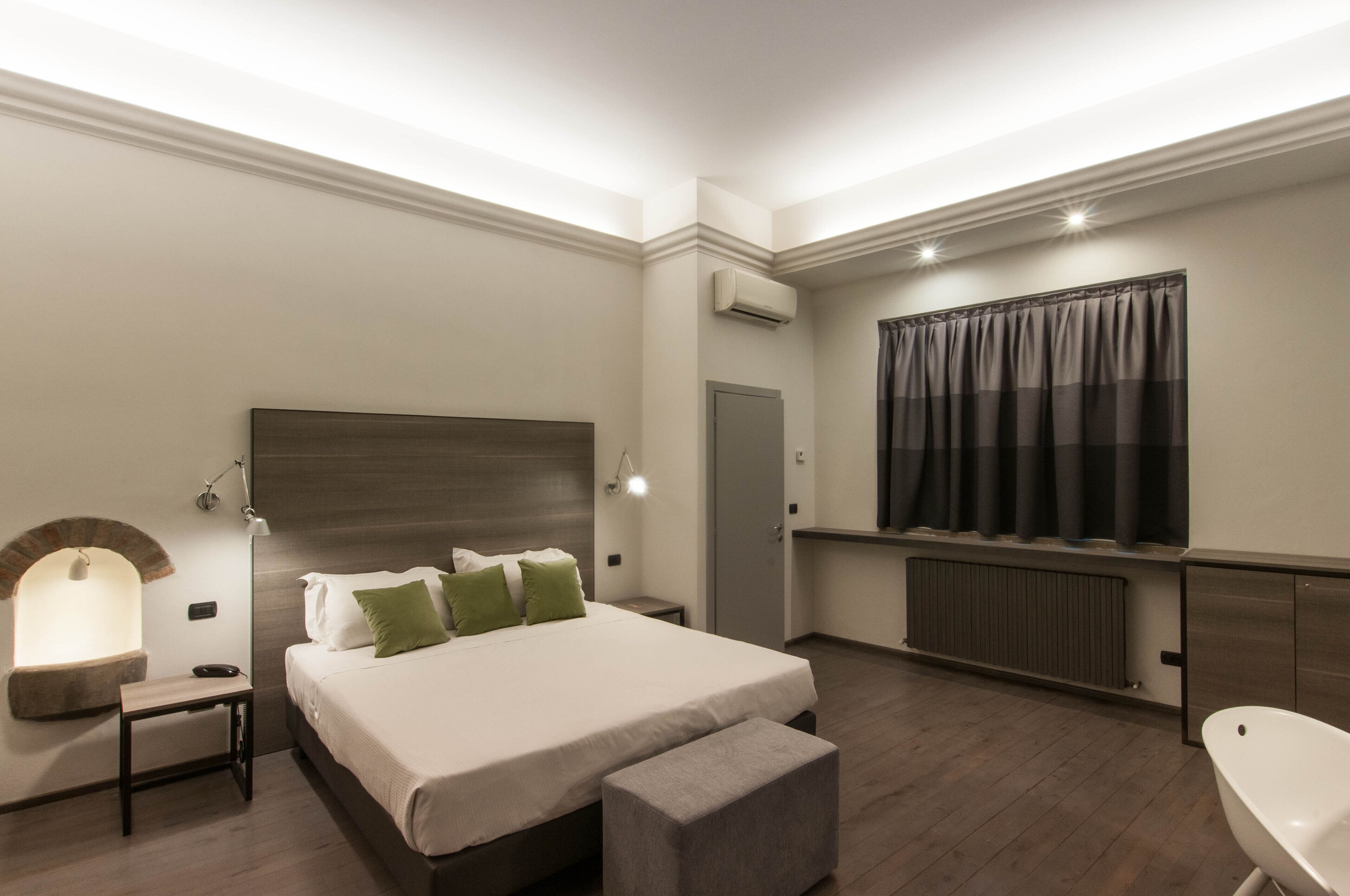 A modern hotel room with a large bed, a bedside table, a bench, a built-in bench at the foot of the bed, a wall-mounted air conditioner, a radiator, a window with curtains, and a wall-mounted light fixture.