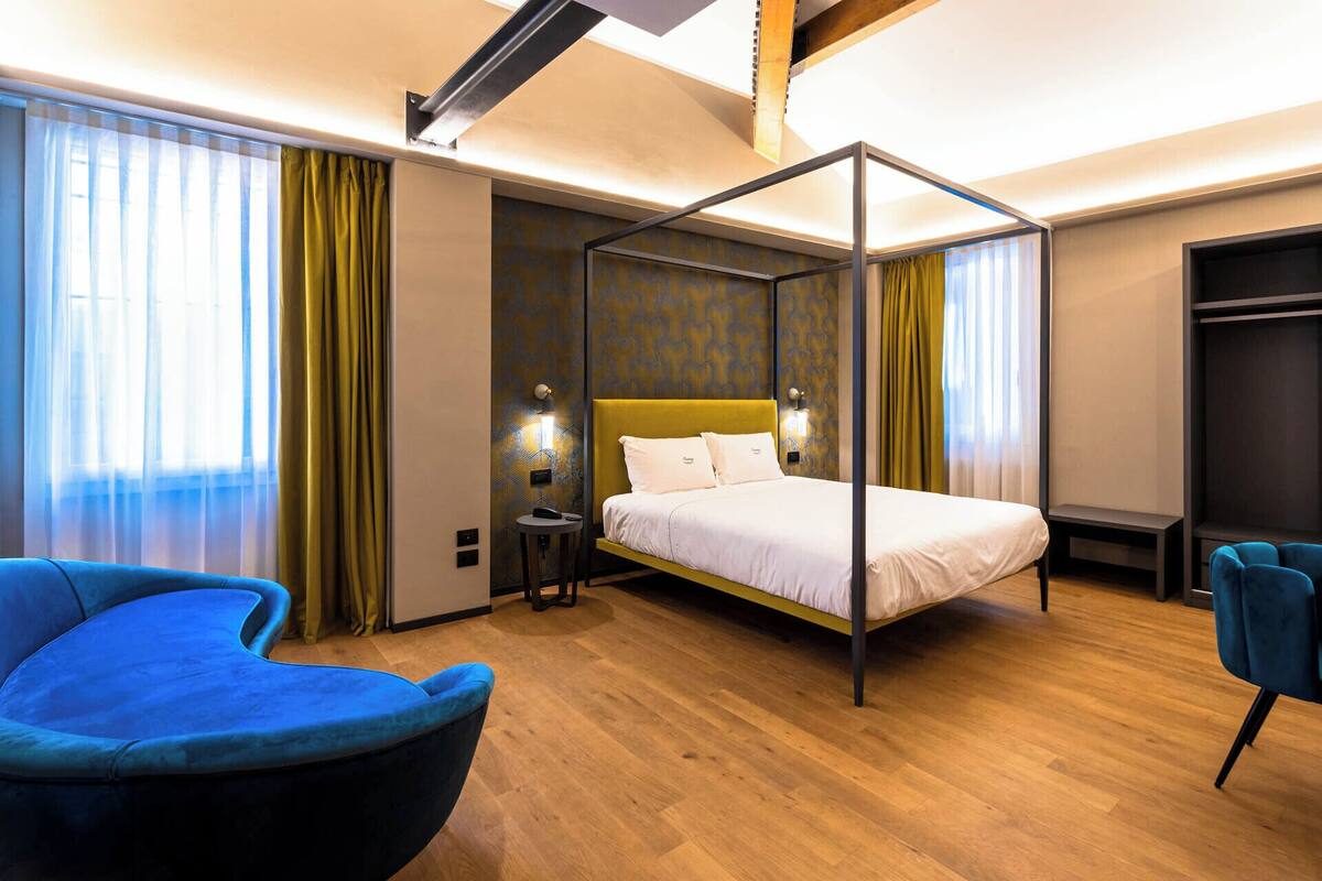 A modern hotel room with a four-poster bed, a blue armchair, a bedside table, and a wardrobe.