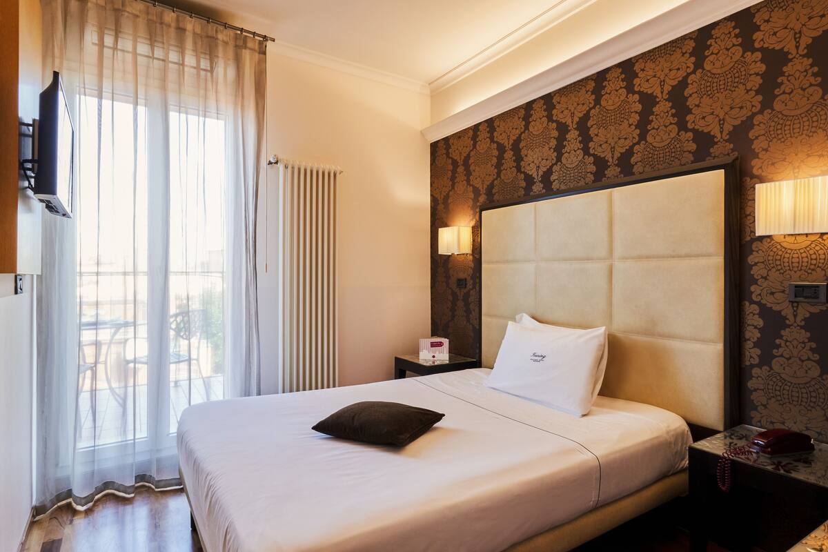 A hotel room with a large bed, a TV mounted on the wall, a bedside table with a lamp, and a view of the outdoors through sheer curtains.
