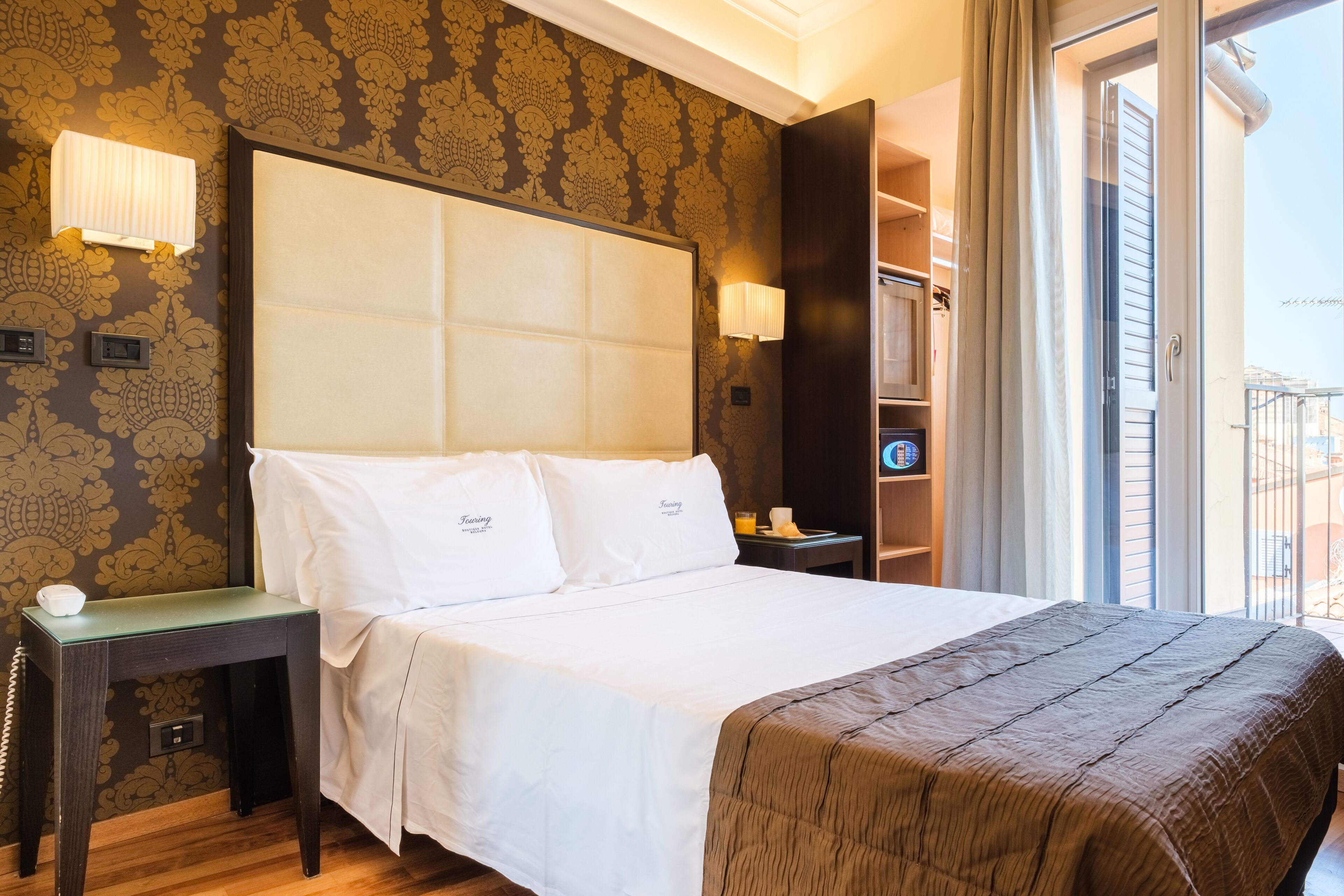 Habitación de hotel con una cama grande, una mesita de noche, una lámpara y vistas de la ciudad a través de la ventana.