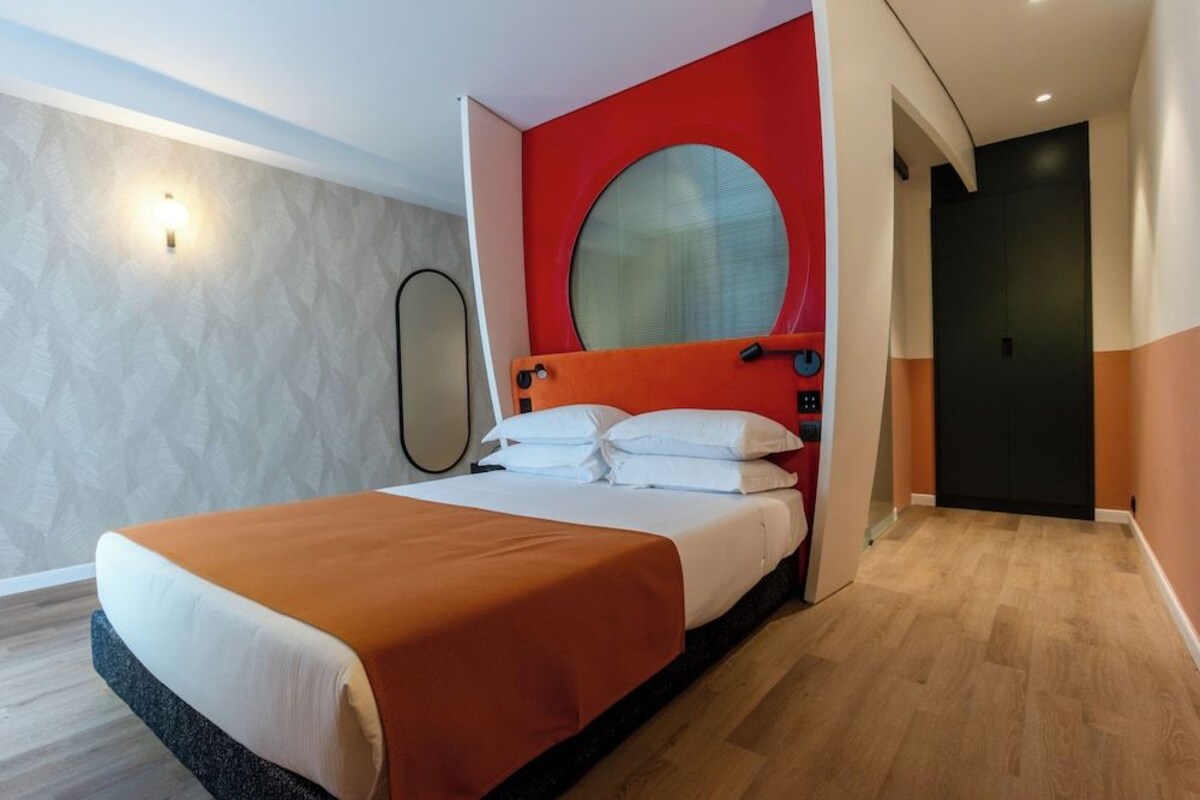 A hotel room with a bed, a round mirror, a red accent wall, and a door.
