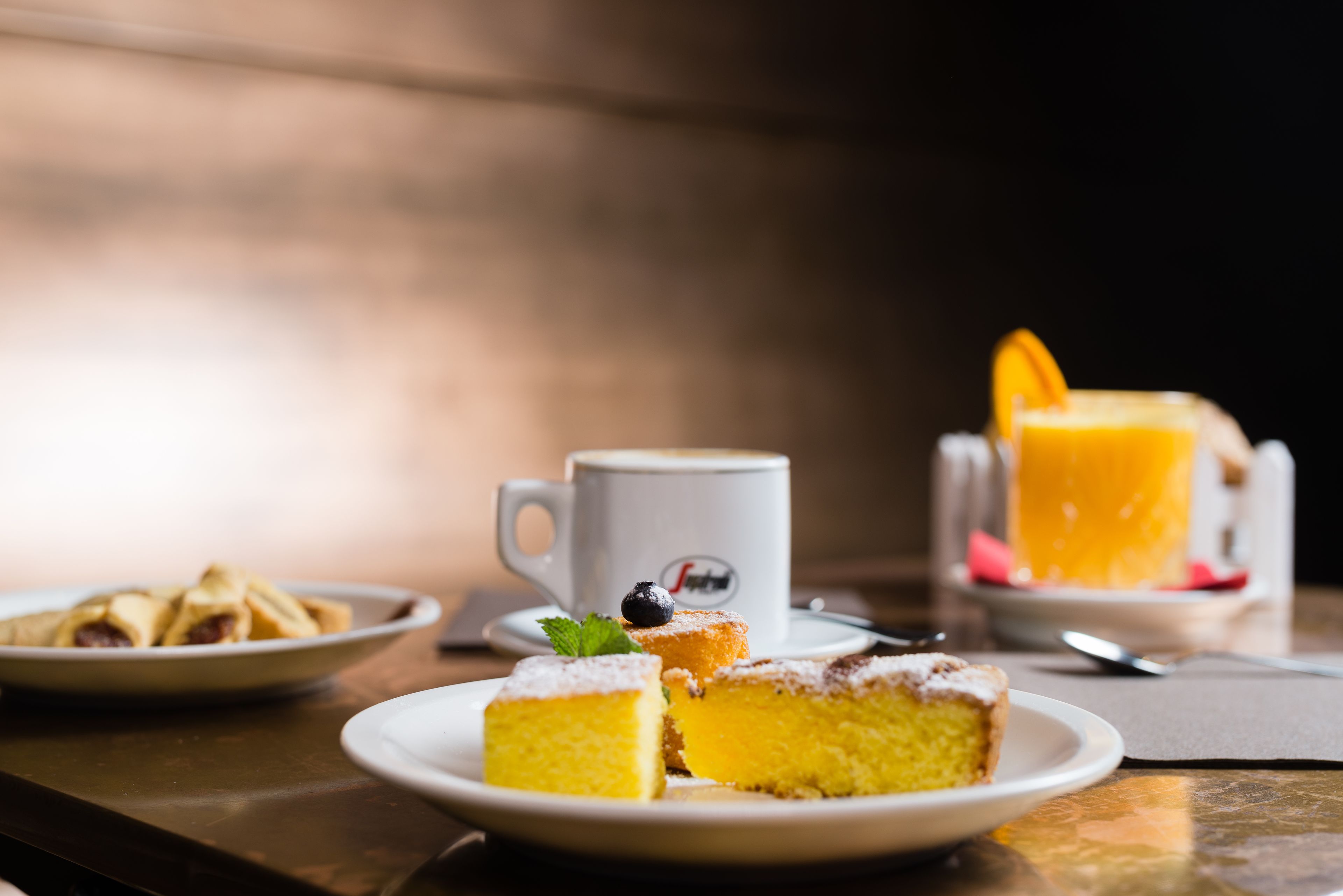 Una mesa con un plato de pastel amarillo, una taza blanca y un vaso de jugo de naranja.