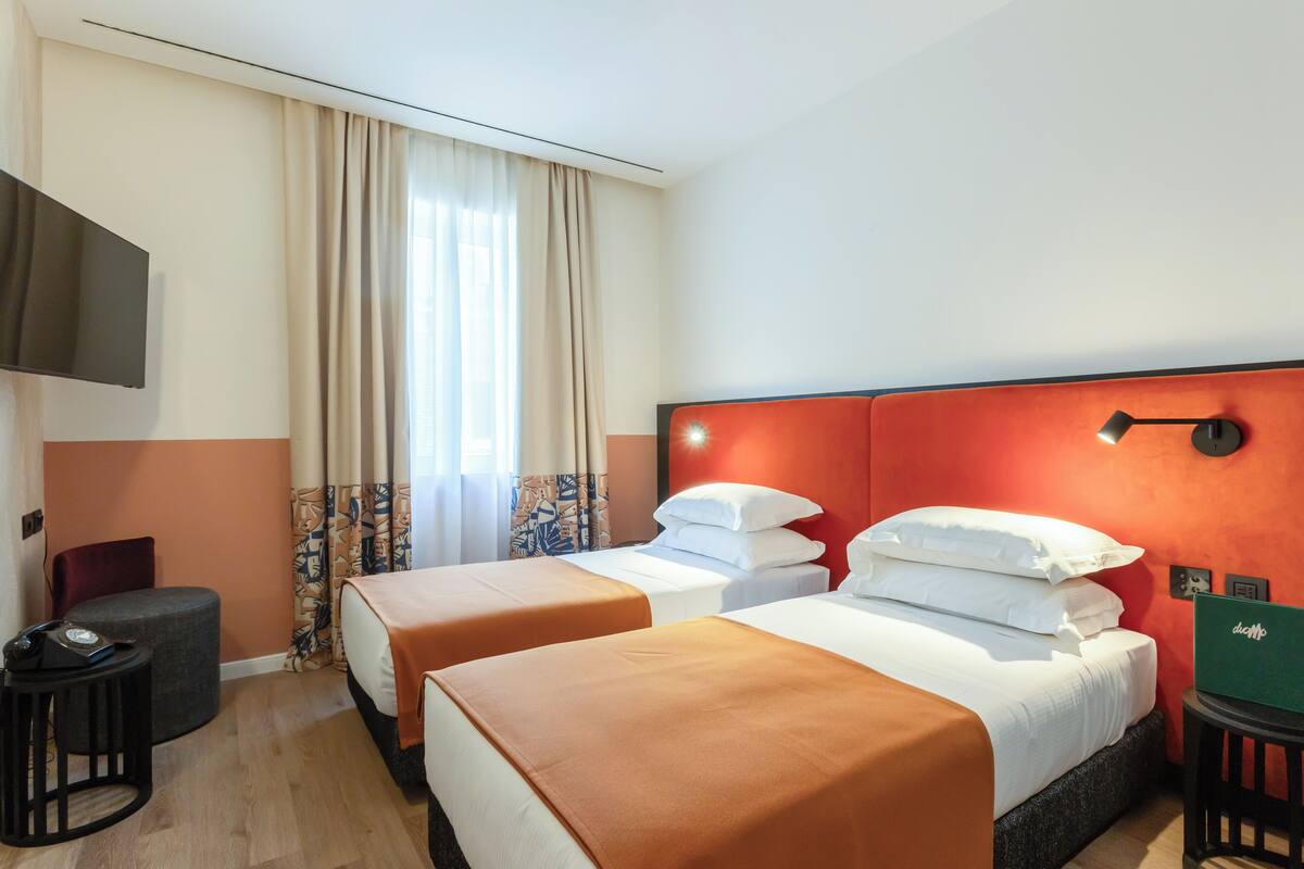 A hotel room with two beds, a TV mounted on the wall, a desk with a chair, and a window with curtains.