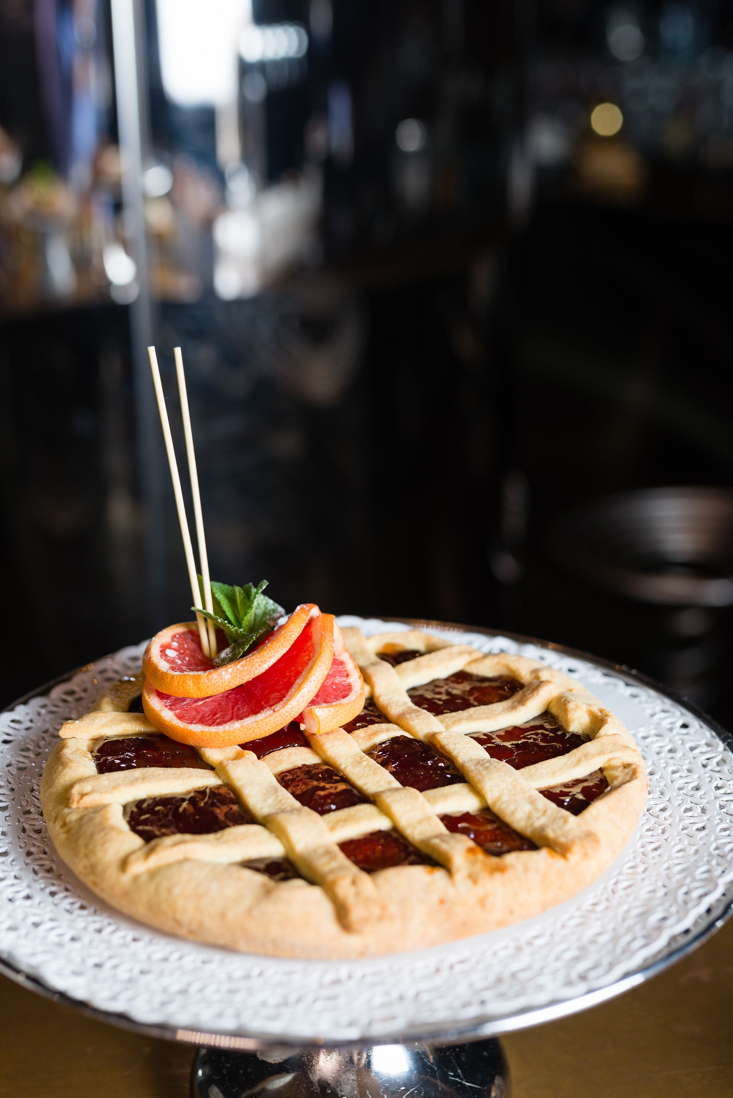 Une tarte recouverte d’une pâte en croisillons, présentée sur une assiette blanche ornée d’un chemin de table, le tout posé sur un présentoir en métal.