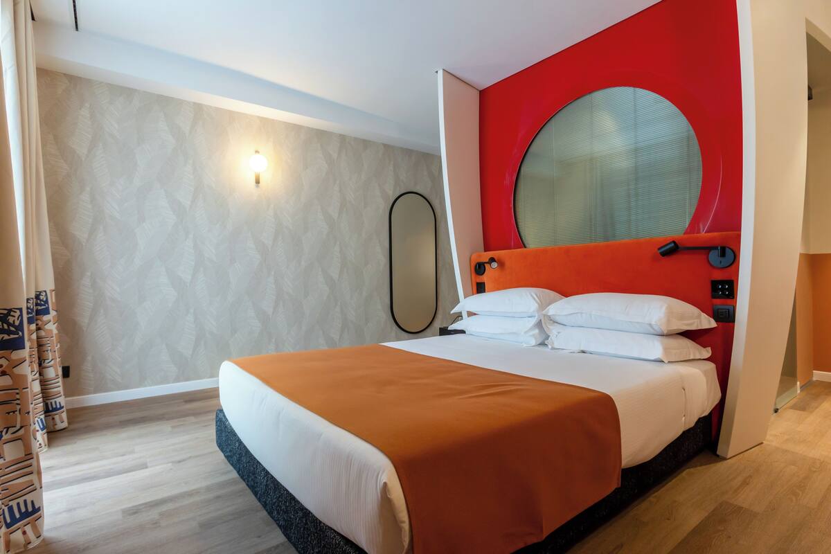 A modern hotel room with a large bed, a red accent wall, and a round mirror.