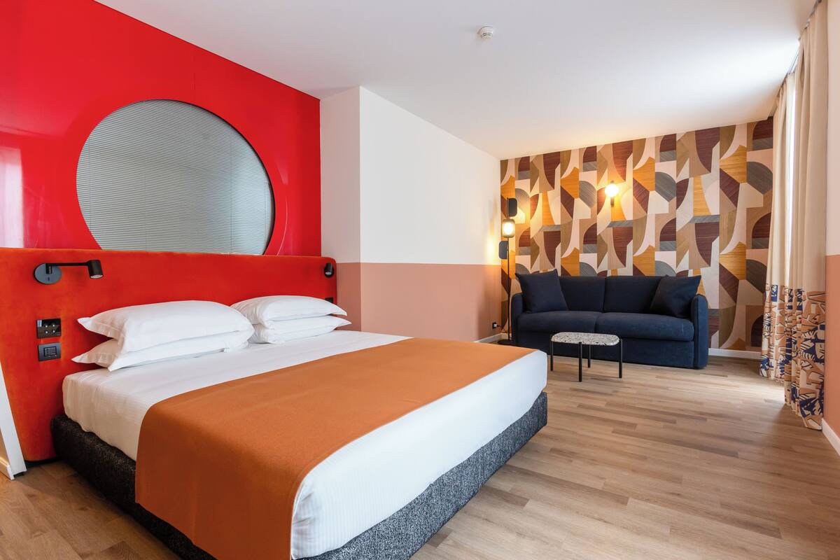 A hotel room with a large bed, a red accent wall, a patterned wall, a blue sofa, and a small table.