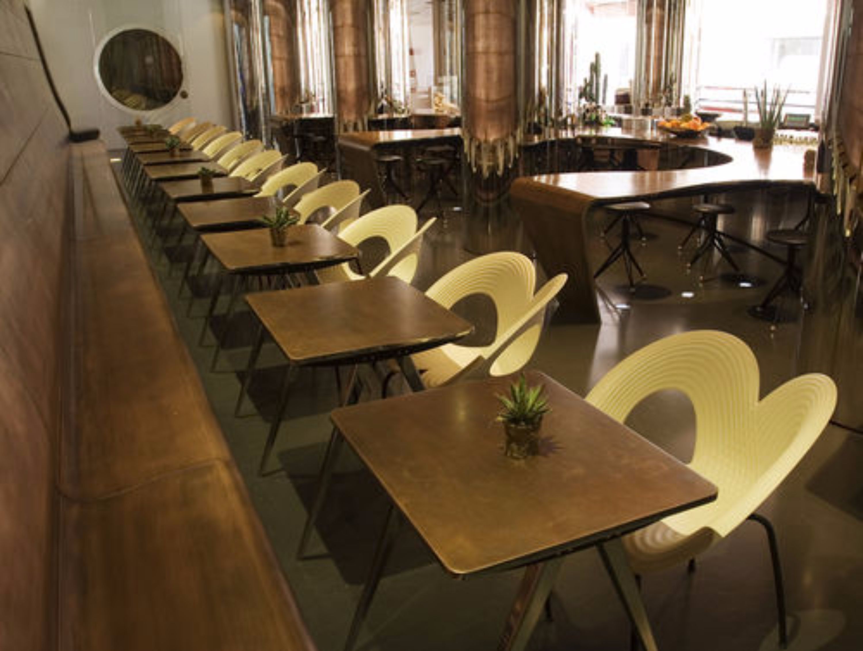 Un comedor amplio con varias mesas y sillas, una zona de bar con taburetes y ventanas grandes que dejan entrar luz natural.