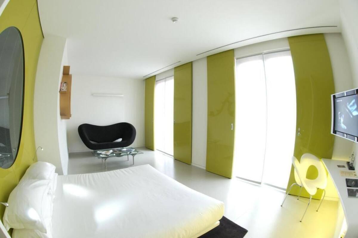 A modern hotel room with a bed, a chair, a desk with a computer, and a TV.
