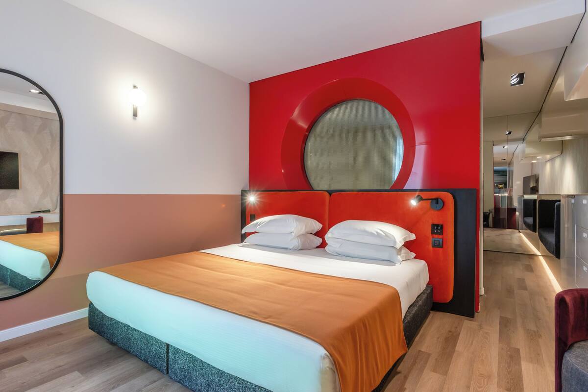 A modern hotel room with a large bed, a round mirror above the bed, and a red accent wall.