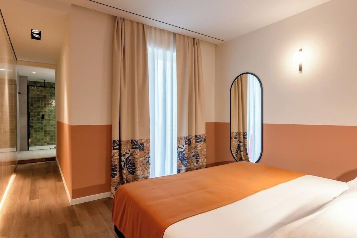 A hotel room with a bed, curtains, a mirror, and a shower area.