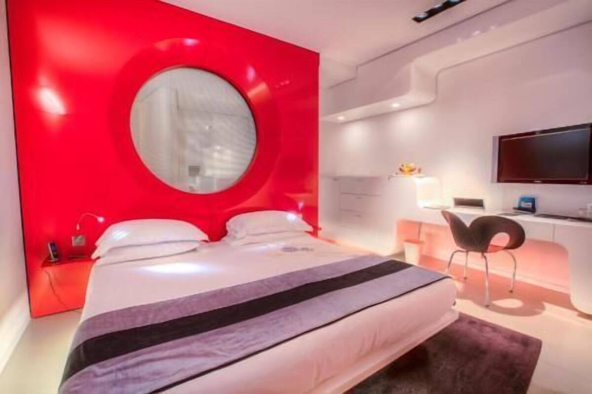 A modern hotel room with a large bed, a round mirror, a desk, and a television.
