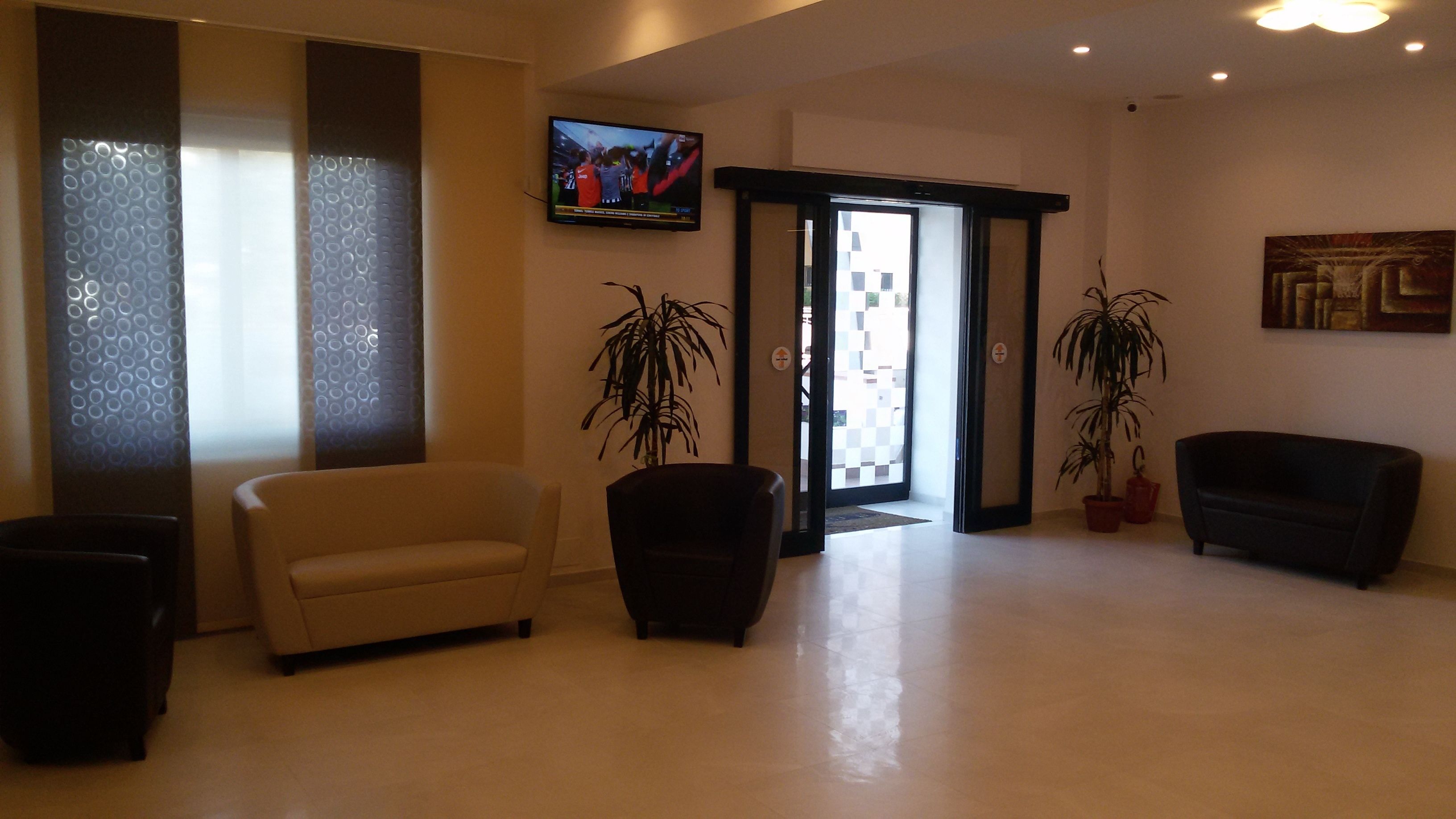 Un hall d’entrée spacieux, avec un canapé beige, un fauteuil noir et deux plantes en pot. Un téléviseur est fixé au mur, et un tableau est visible sur la droite.