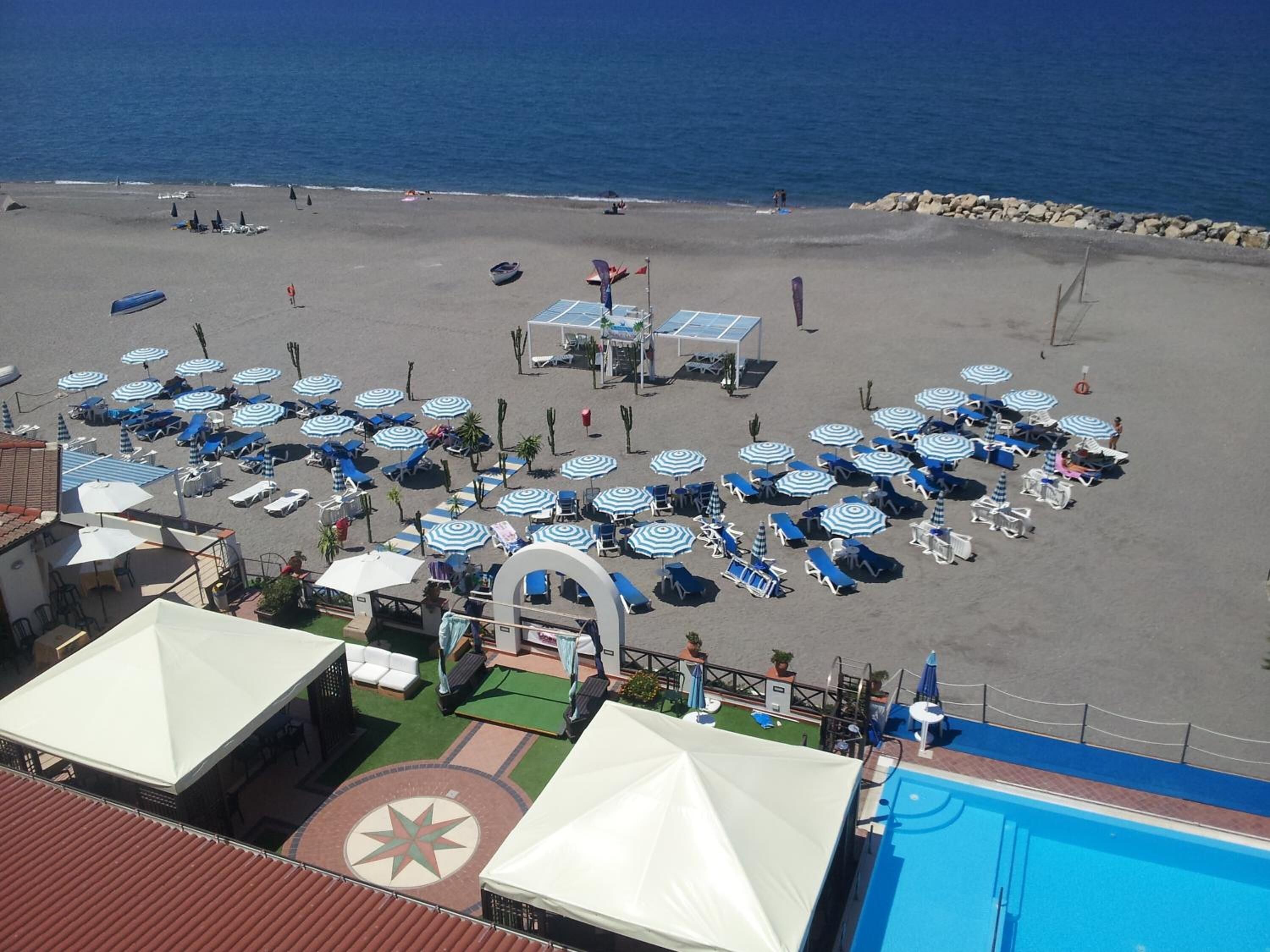 Un hotel frente a la playa con numerosas tumbonas y sombrillas, piscina y vistas al mar.