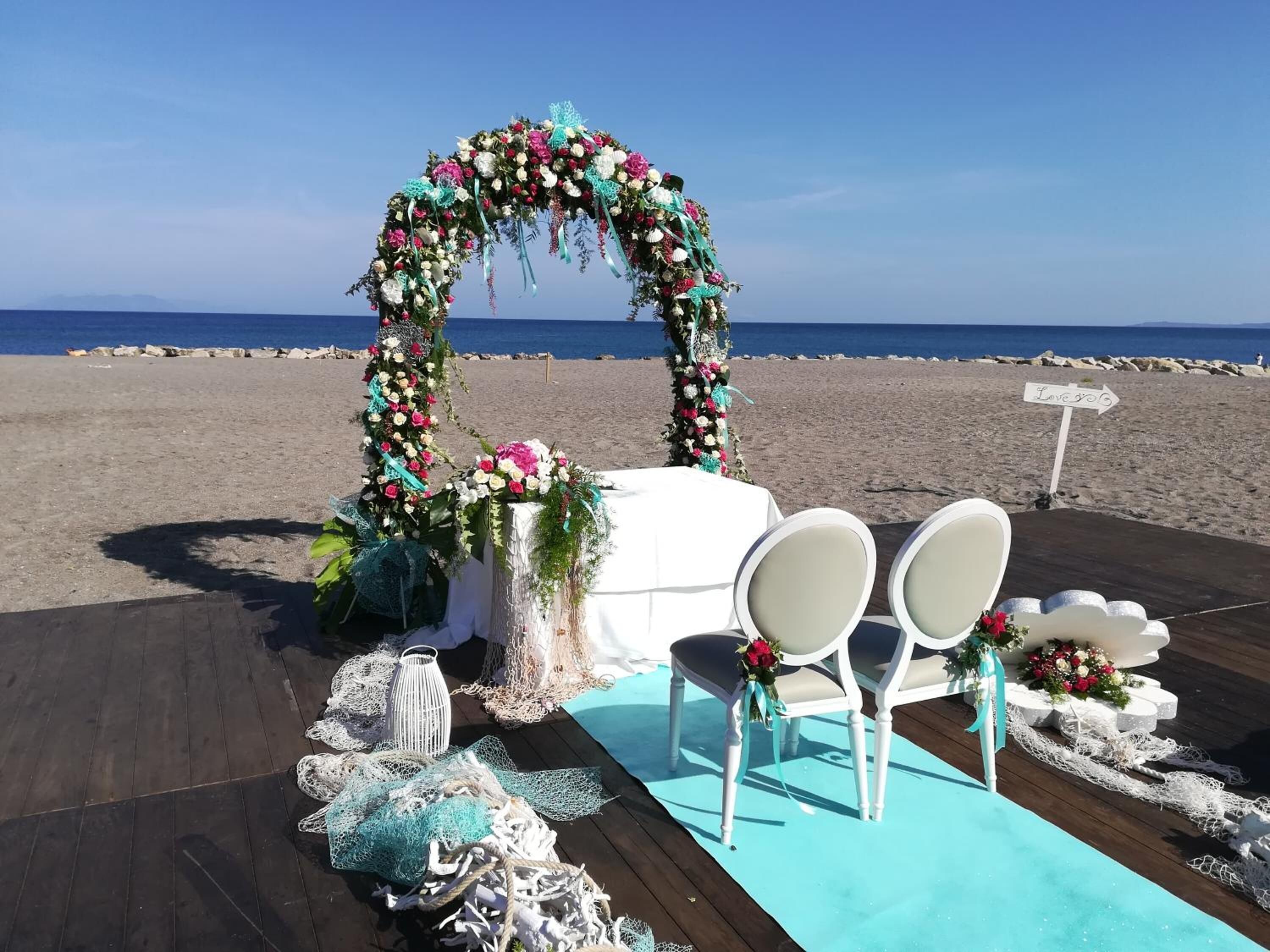 A beach wedding setup with a floral arch, white chairs, and a sign that says "Love is the answer".