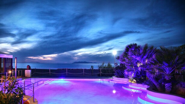 A swimming pool with a curved edge, surrounded by palm trees and a view of the ocean at dusk. - Hotel Il Gabbiano Beach (Terme Vigliatore)